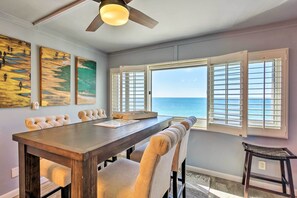 A dining area with a long wooden table and cushioned chairs, large windows with shutters, and artwork on the walls. - PCB Beachfront 'coastal Abode' at Pinnacle Port! (Panama City Beach)