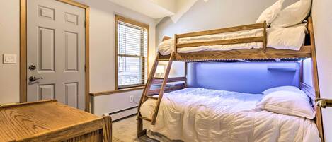 Uma cama de beliche com roupa de cama branca, uma escada de madeira, uma mesinha de madeira e uma janela com persianas.