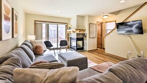 A modern living room with a grey sofa, ottoman, and a fireplace. There is a TV mounted on the wall and a staircase leading to another level. - Winter Park Townhome w/ Hot Tub, Ski Shuttle! (Winter Park)