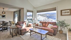 A living room with a large window offering a view of a snowy mountain landscape, a brown leather sofa, a wooden coffee table, and a dining area with a table and chairs. - Walkable Winter Park Condo ~ 4 Mi to Slopes! (Winter Park)