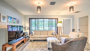 Ein Wohnzimmer mit einer Couch, einer Bank und einem Sessel. Zwei Pendelleuchten, ein Fenster mit Jalousien und eine Tür sind vorhanden.