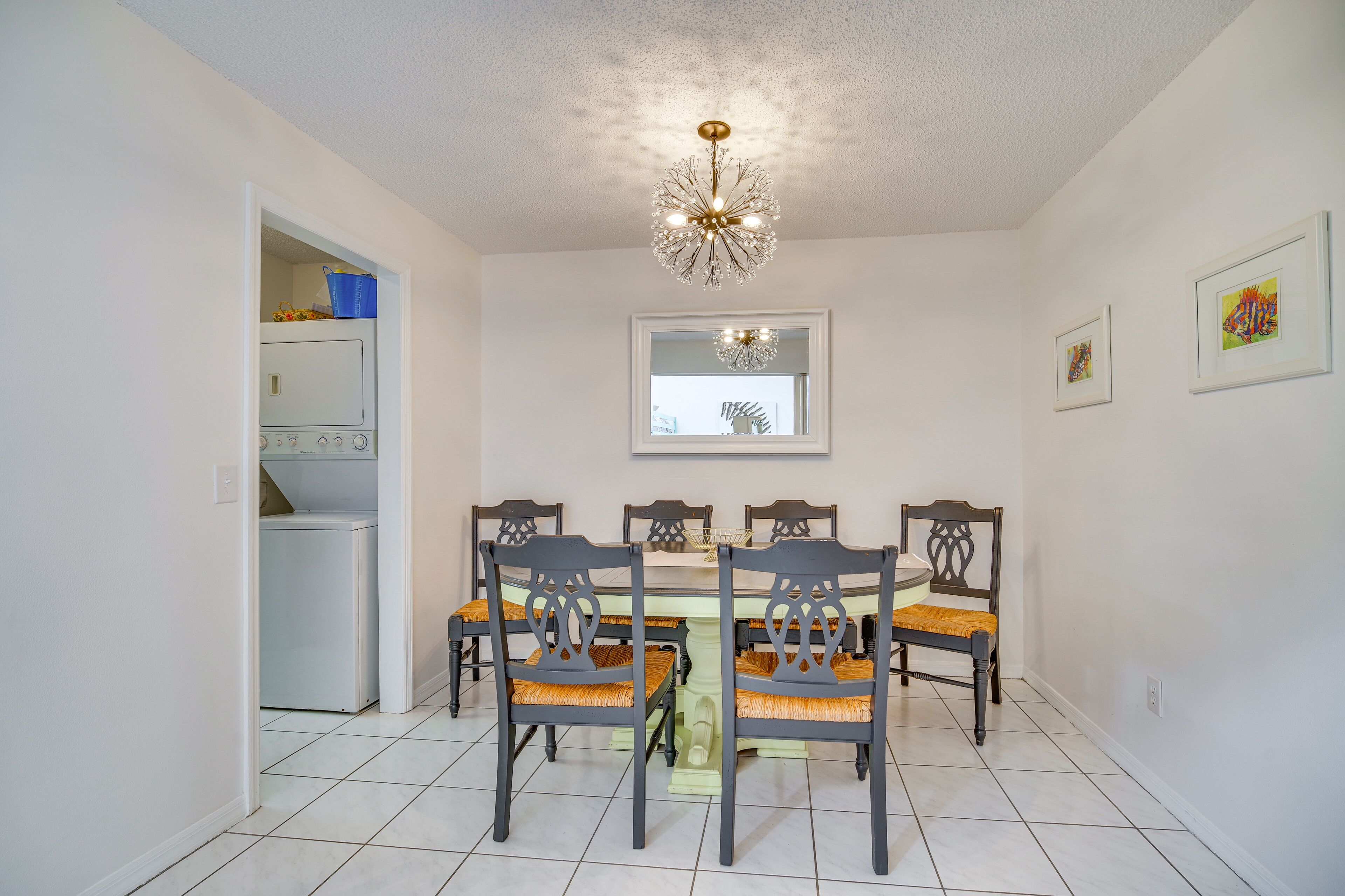 A dining area with a round table, six chairs, and a chandelier. There is a laundry room to the left and a mirror on the wall.