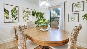 A round wooden dining table with wicker chairs, surrounded by framed tropical plant prints on the walls.