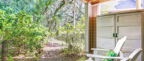 A covered porch with white chairs and a green patterned rug, surrounded by lush tropical vegetation.