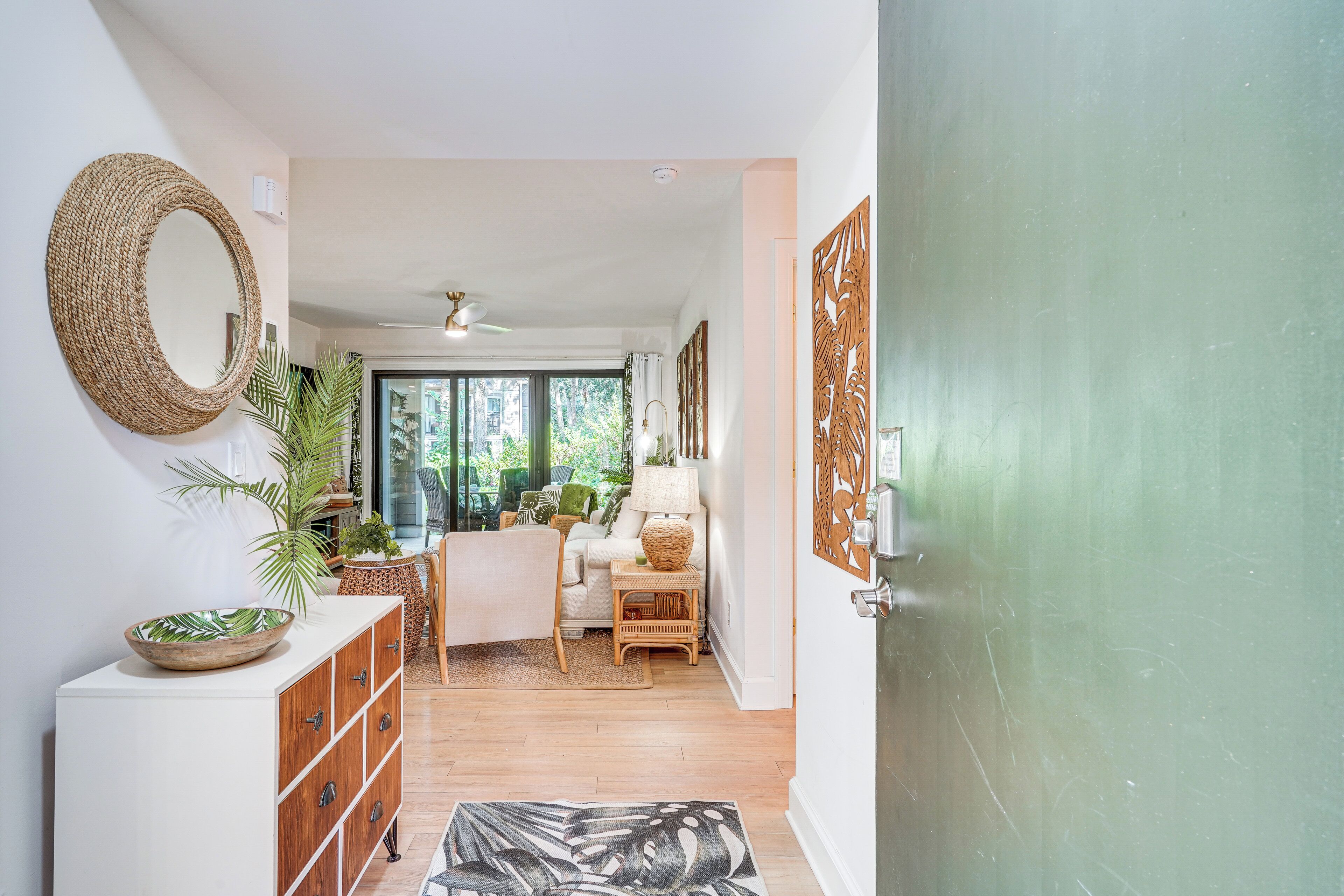 A hallway with a white cabinet, a woven mirror, a large woven bowl, a black and white abstract rug, a wicker chair, a wicker side table, and a green door.