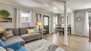 Un salon moderne avec un canapé d’angle gris, un fauteuil bleu et un tapis à motifs. On aperçoit à l’arrière-plan un coin repas avec une table et des chaises.