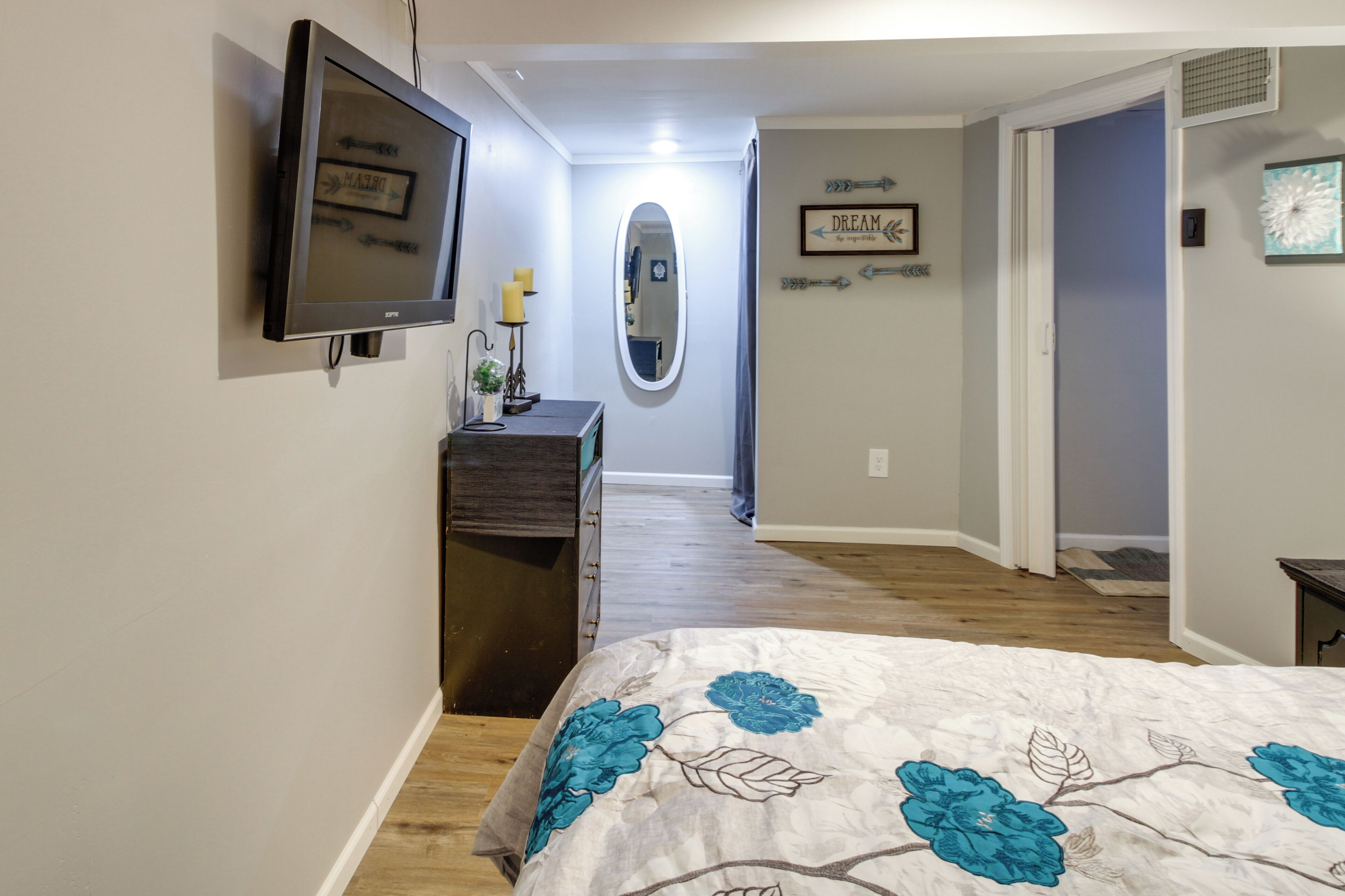 A bedroom with a bed, a dresser, a television mounted on the wall, a mirror, and a door leading to another room.