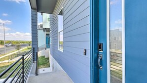 Ein modernes Gebäude mit blauer Fassade, einem überdachten Außenbereich mit Metallgeländer und freiem Blick auf eine Straße und Grünflächen im Hintergrund.