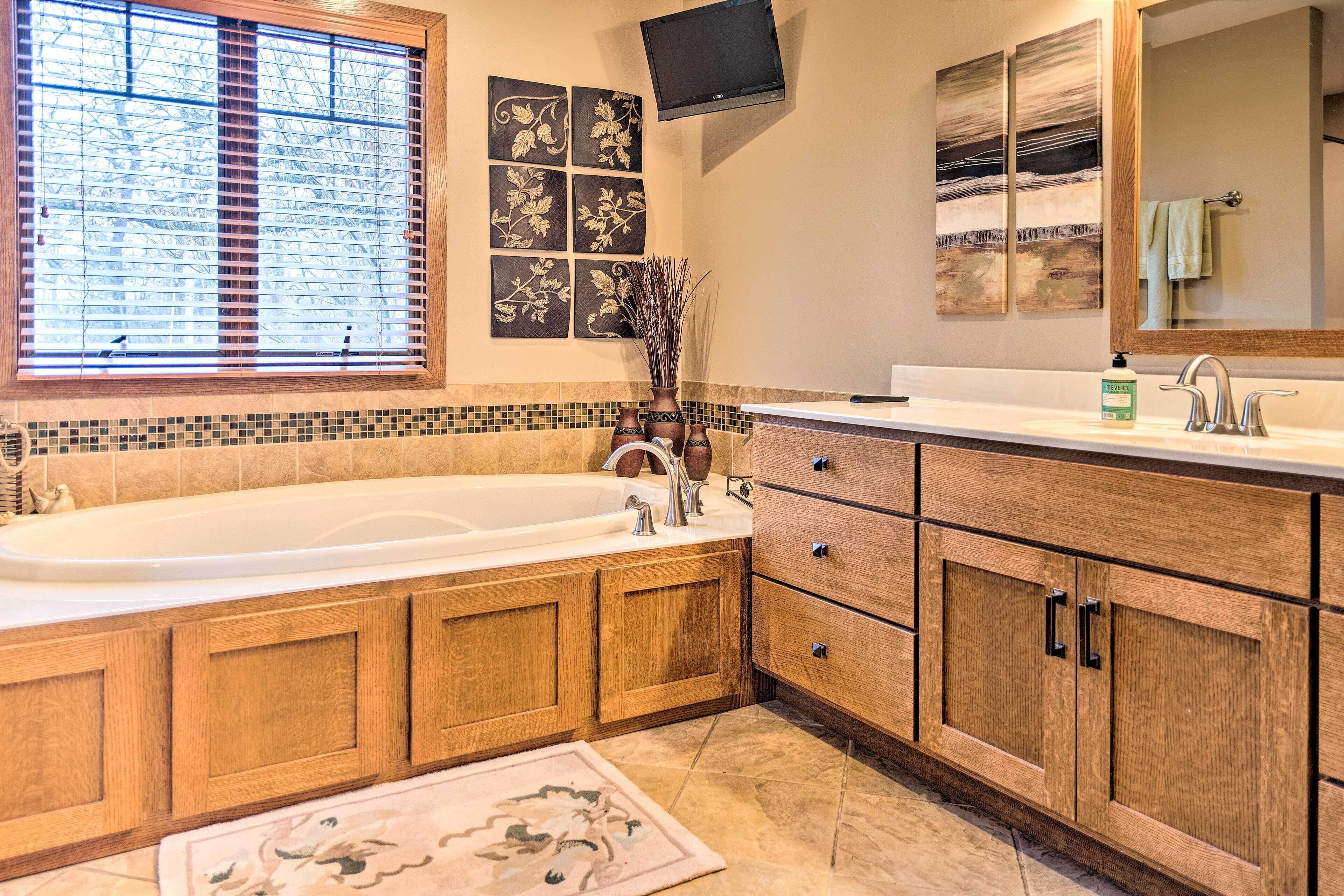 A modern bathroom with a large bathtub, wooden cabinetry, a tiled backsplash, and a flat-screen TV mounted above the sink.