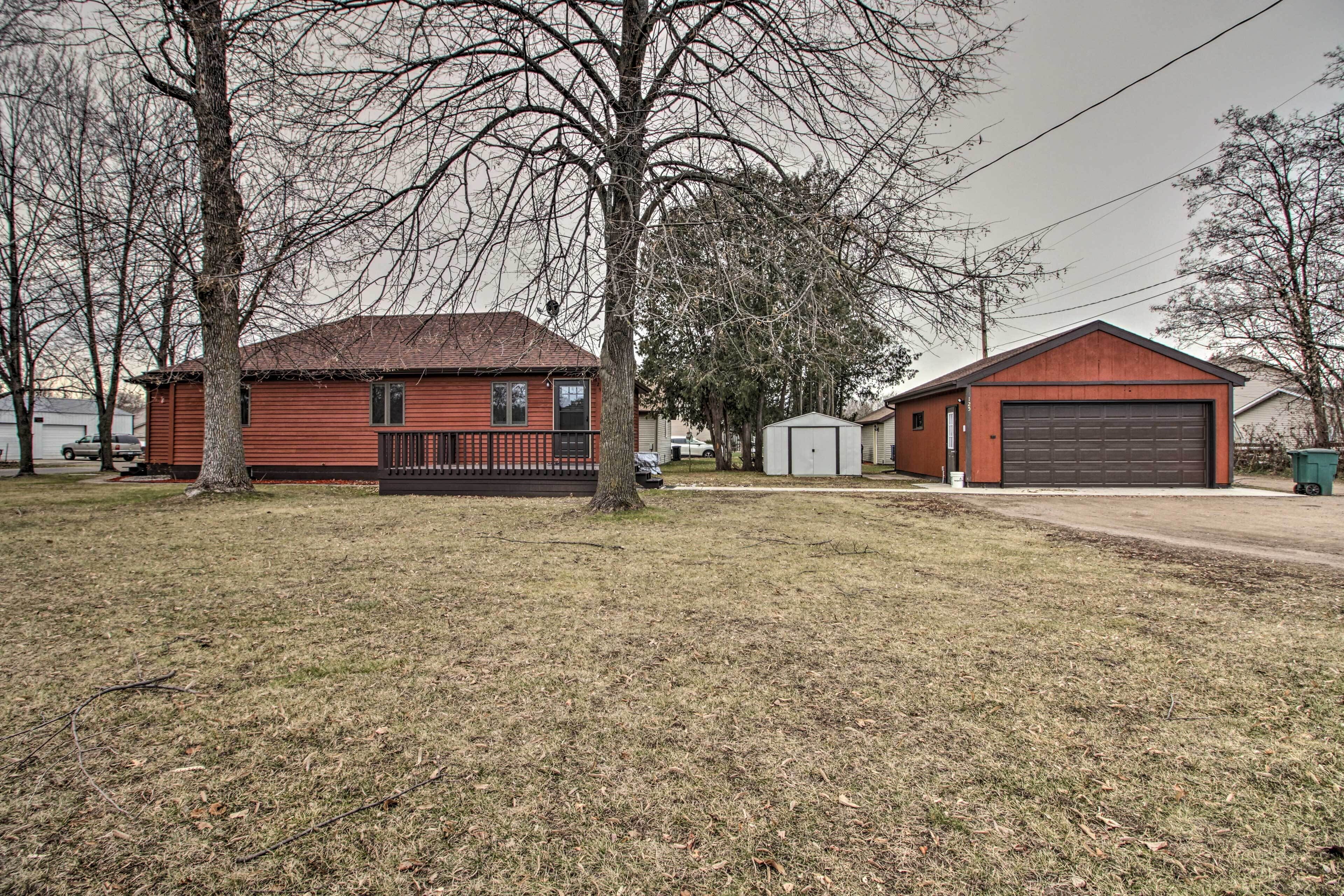A red single-story house with a garage, a small shed, and a tree in front.