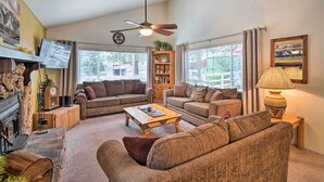 A spacious living room with two sofas, a coffee table, a fireplace, a ceiling fan, and large windows with curtains. - 3 Mi to Skiing: Big Bear Cabin w/ Deck (Big Bear City)