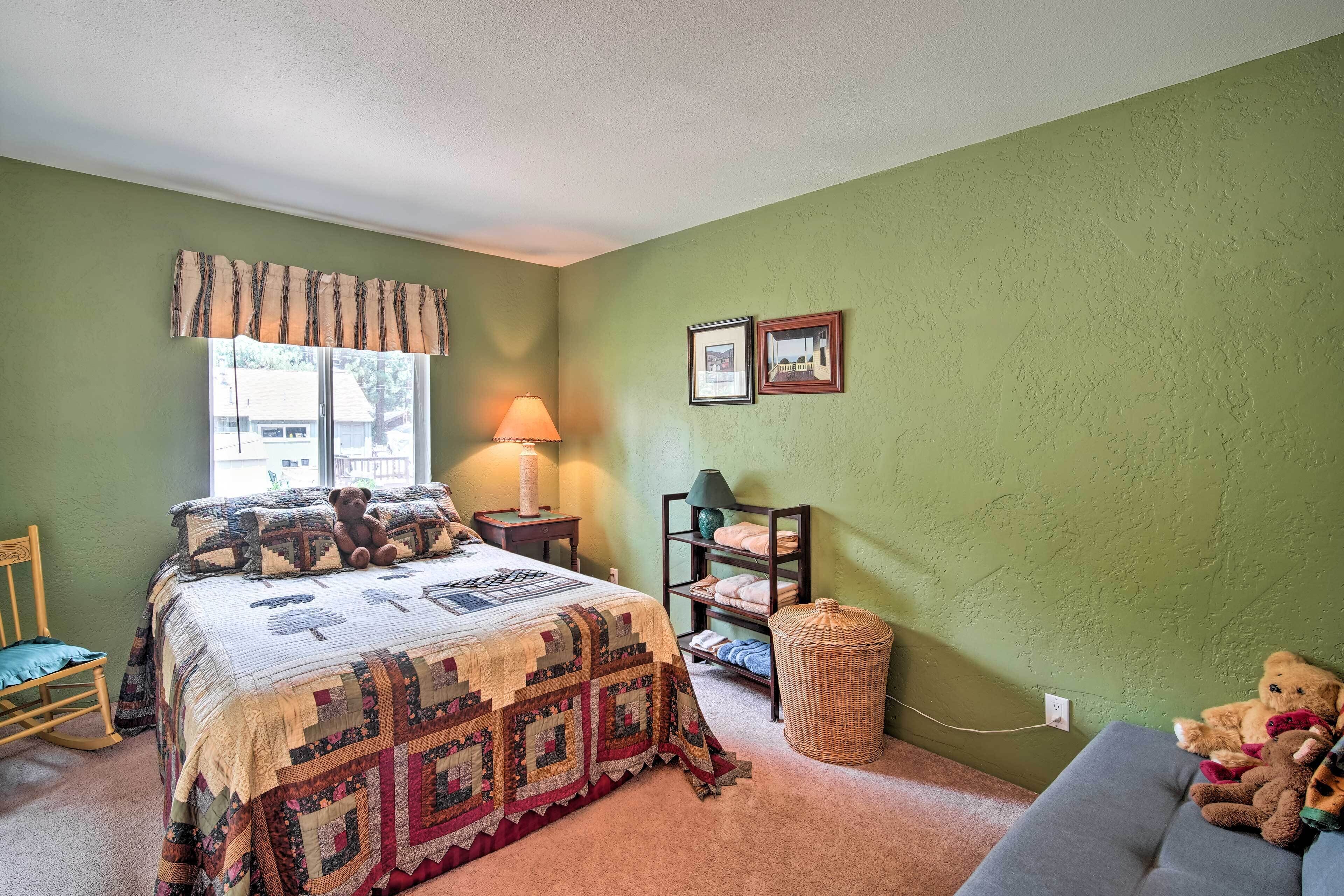 A bedroom with a large bed, a nightstand, a lamp, a chair, a shelf with folded clothes, and a teddy bear on a couch.