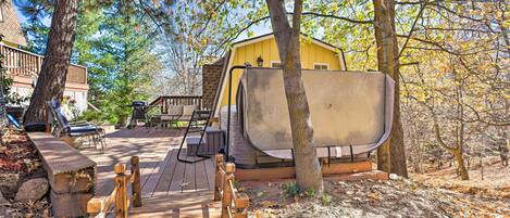 A wooden deck leading to a yellow cabin with a large window, surrounded by trees and a stone wall.