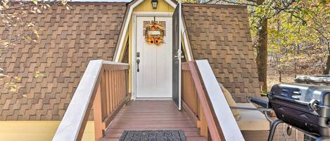 A wooden deck leading to a white door with a wreath, a brick wall, and a barbecue grill in the background.