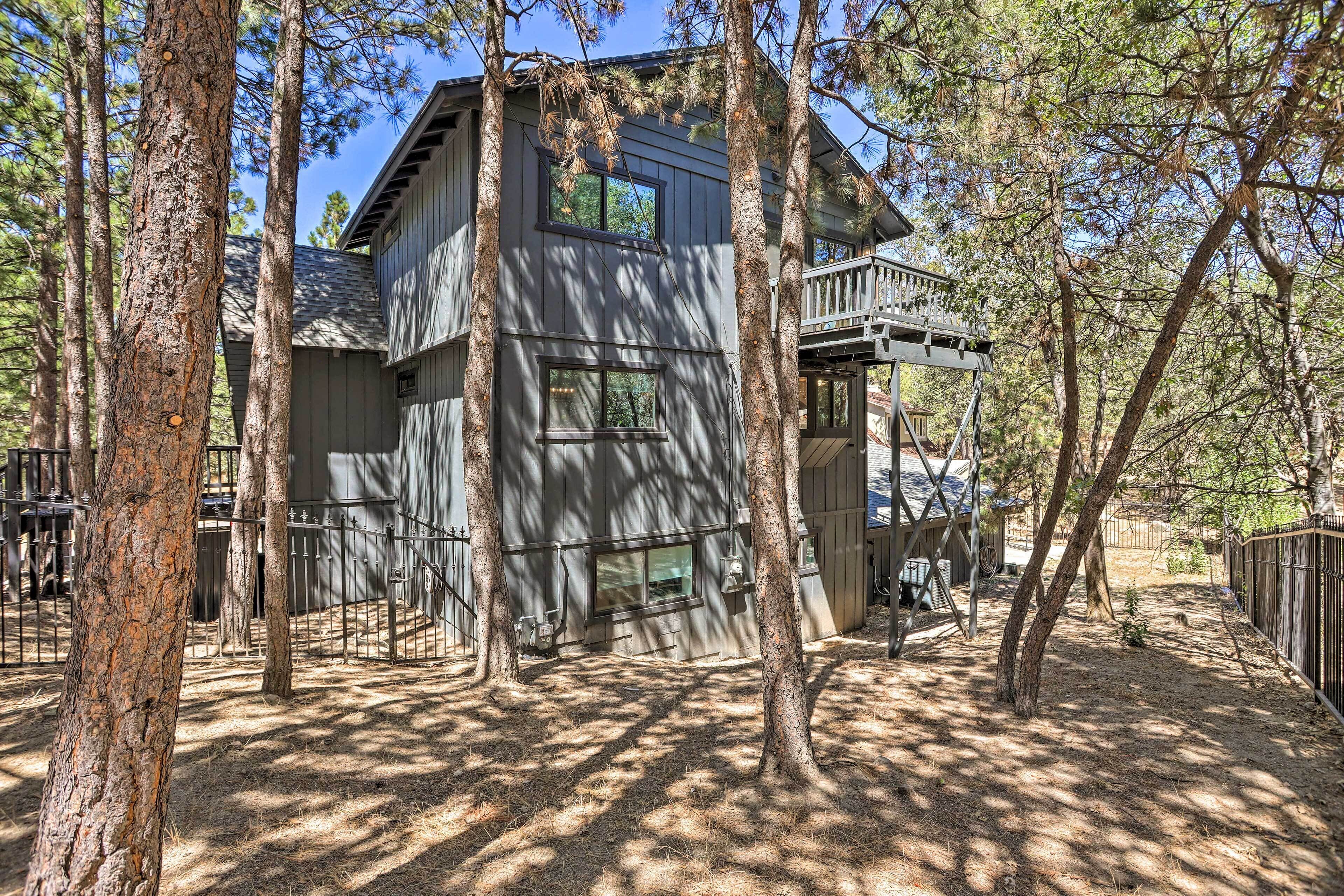 A two-story cabin with a deck, surrounded by tall trees and a fenced yard.