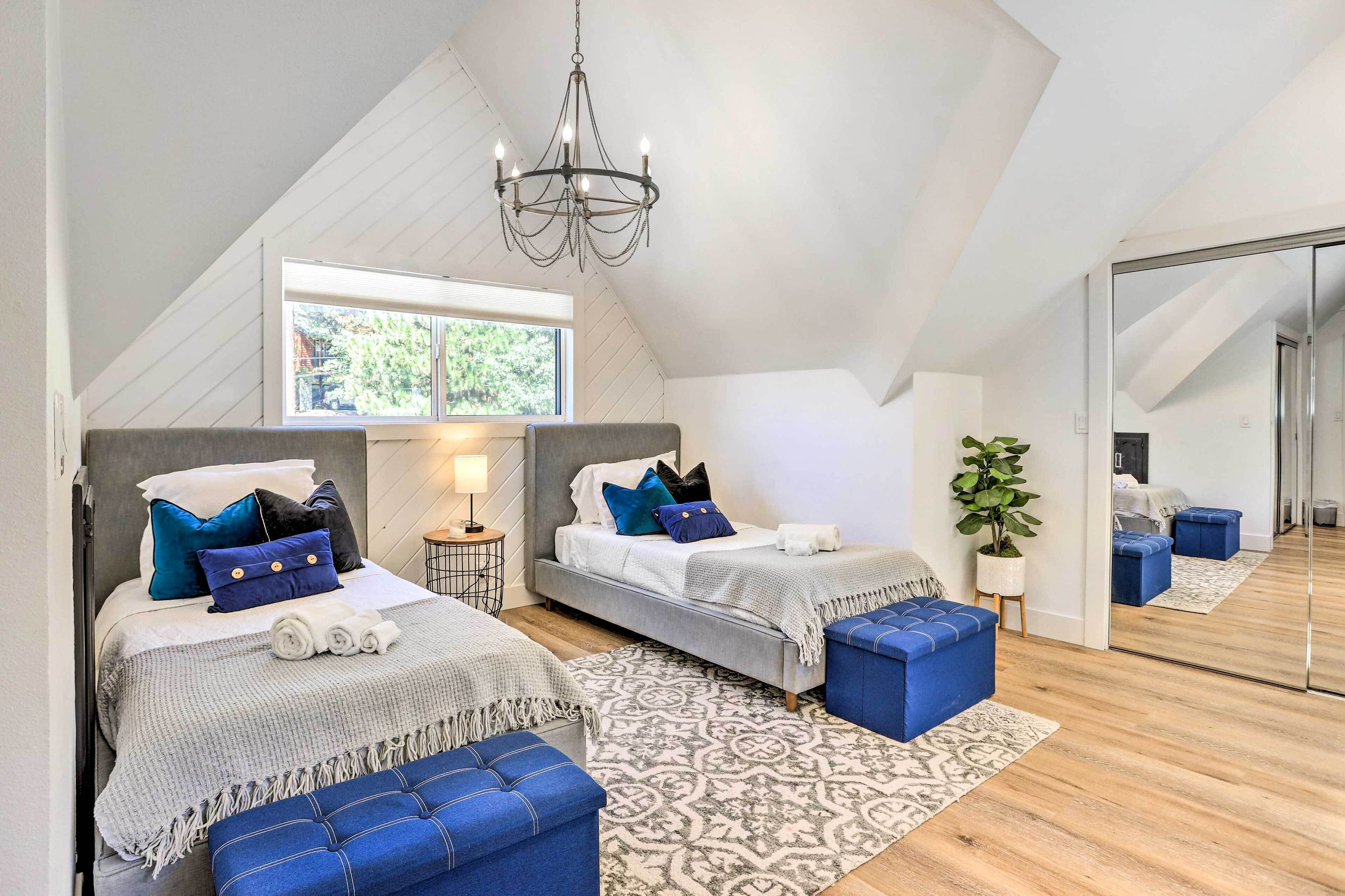 Un dormitorio moderno con techo inclinado, dos camas con ropa de cama azul y blanca a juego, una araña y un espejo grande que refleja la habitación.