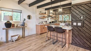 A modern kitchen with wooden cabinets, a central island with bar stools, pendant lighting, and a herringbone-patterned wall. - Luxe Timberland Villa w/ Lake Arrowhead Access! (Lake Arrowhead)