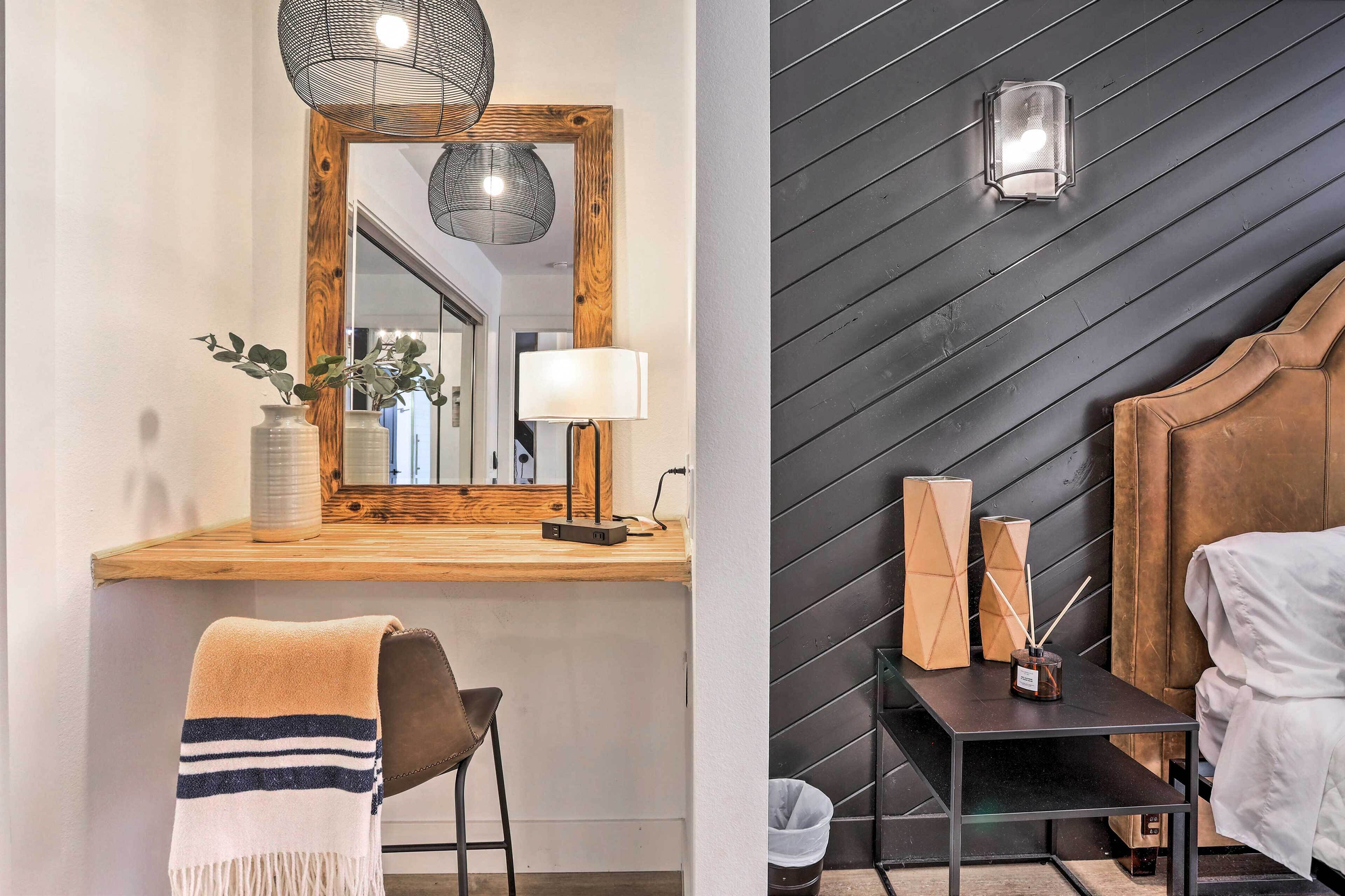 A modern hotel room with a wooden desk, a mirror, a lamp, a bed, and a wall-mounted light fixture.