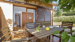 Een terras buiten met houten meubels, een tafel met bekers en glazen, en uitzicht op een tuin en een gebouw ingang.