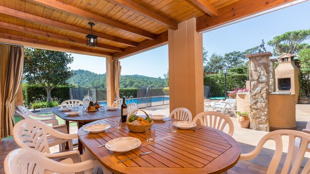 An outdoor wooden table set with plates, glasses, and a basket of fruit, surrounded by white chairs, with a pool and greenery in the background. - 2CIP01-10pax - House with capacity for 10 people and private pool located in a quiet area (Vidreres)