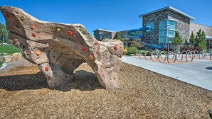 A rock climbing wall with various holds, a paved walkway, and a building with a stone facade in the background.
