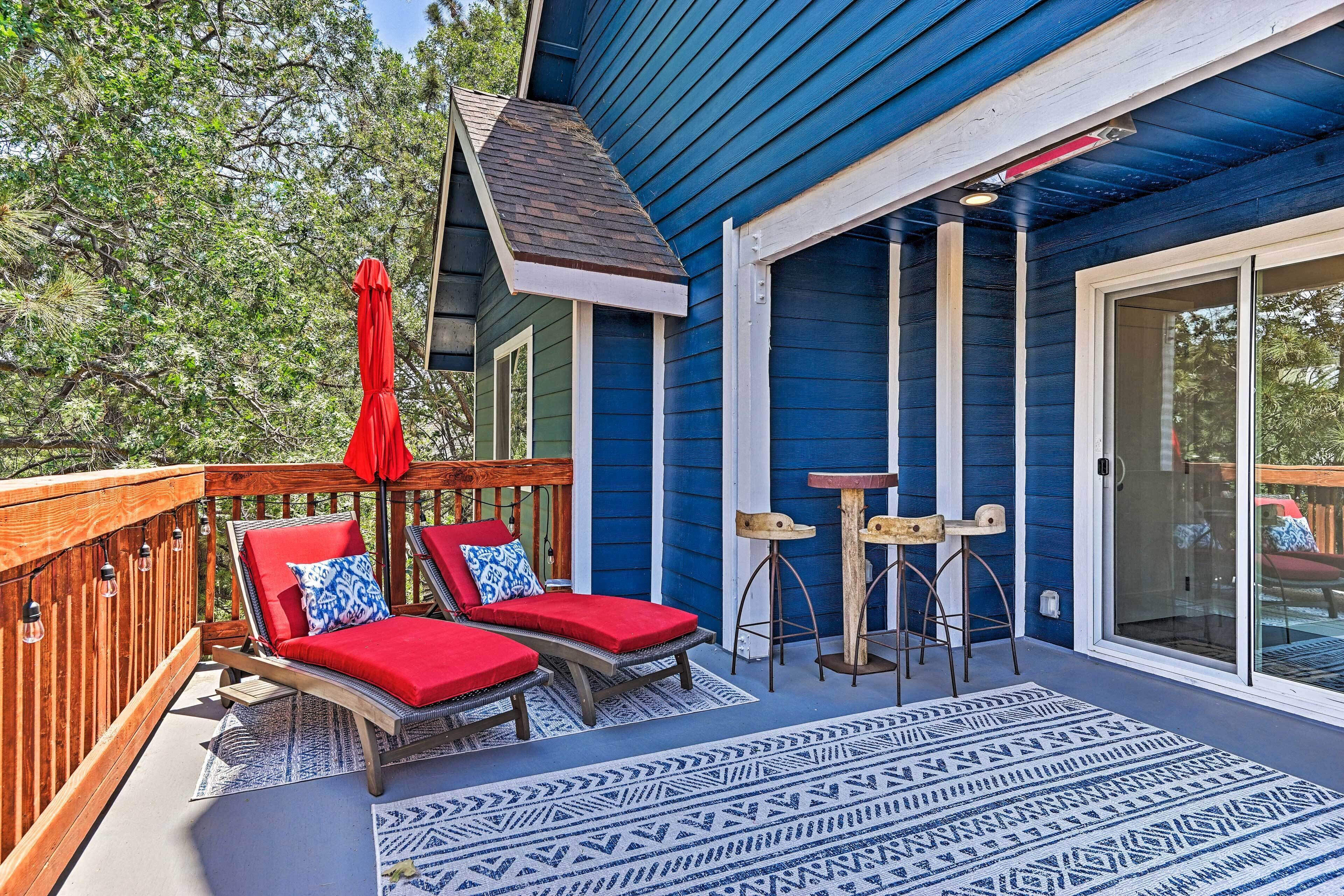 A blue house with a wooden deck, two red lounge chairs, a red umbrella, and a patterned rug.