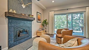 A living room with a stone fireplace, leather sofas, and a large window with a view of trees. - Luxe Lake Arrowhead Farmhouse w/ Deck + Patio (Lake Arrowhead)