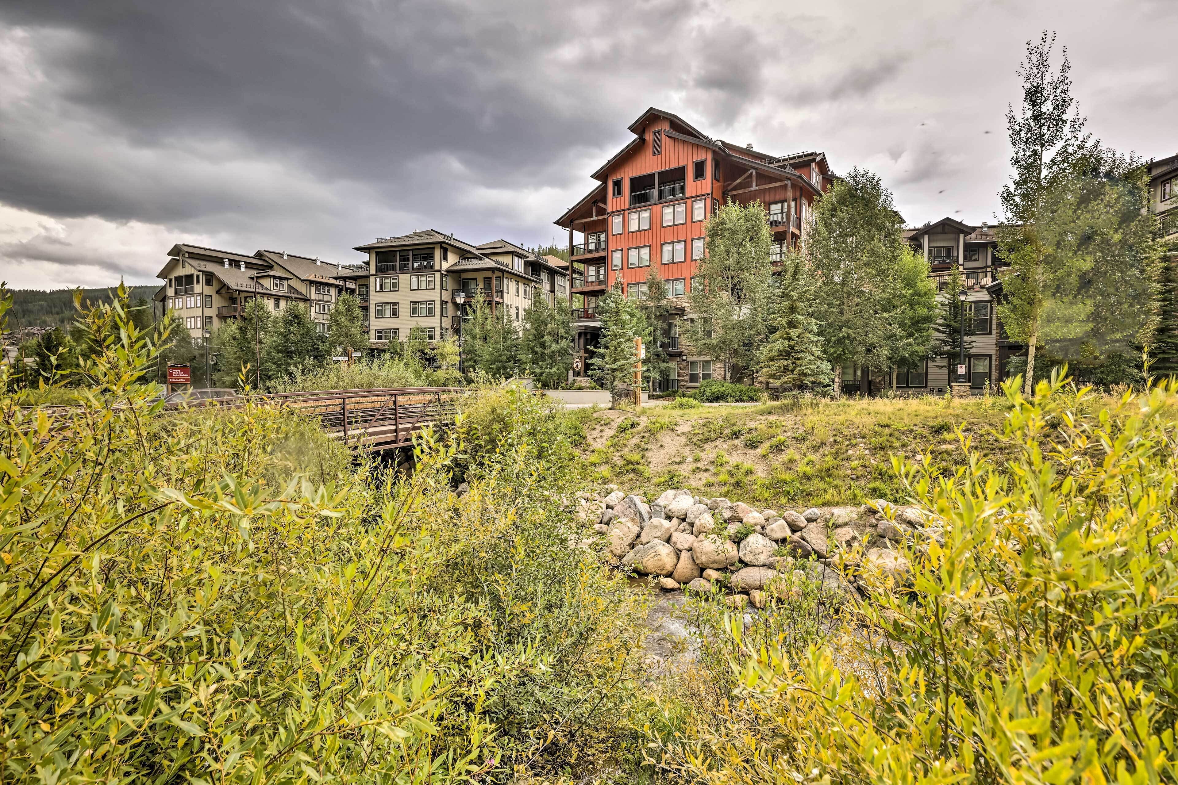 Un immeuble à plusieurs étages, dont l’architecture combine des éléments modernes et traditionnels, entouré d’une végétation luxuriante et traversé par un petit ruisseau.