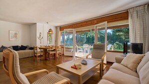 Ein geräumiges Wohnzimmer mit einer Couch, Sesseln, einem Couchtisch und einem Fernseher. Es gibt einen Essbereich mit Tisch und Stühlen, sowie große Fenster mit Blick auf Grünflächen.