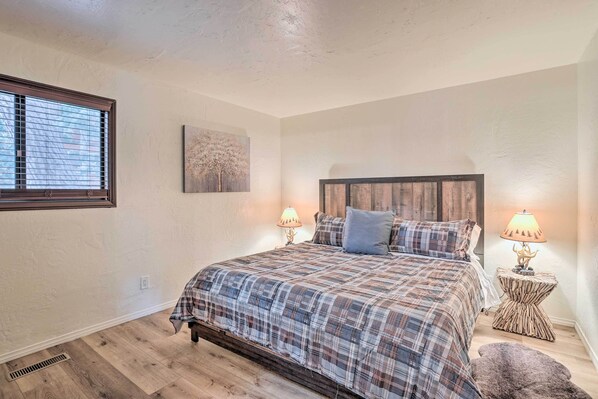A bedroom with a plaid bedspread, two bedside lamps, a headboard, a window with blinds, a small rug, and a wall-mounted artwork.