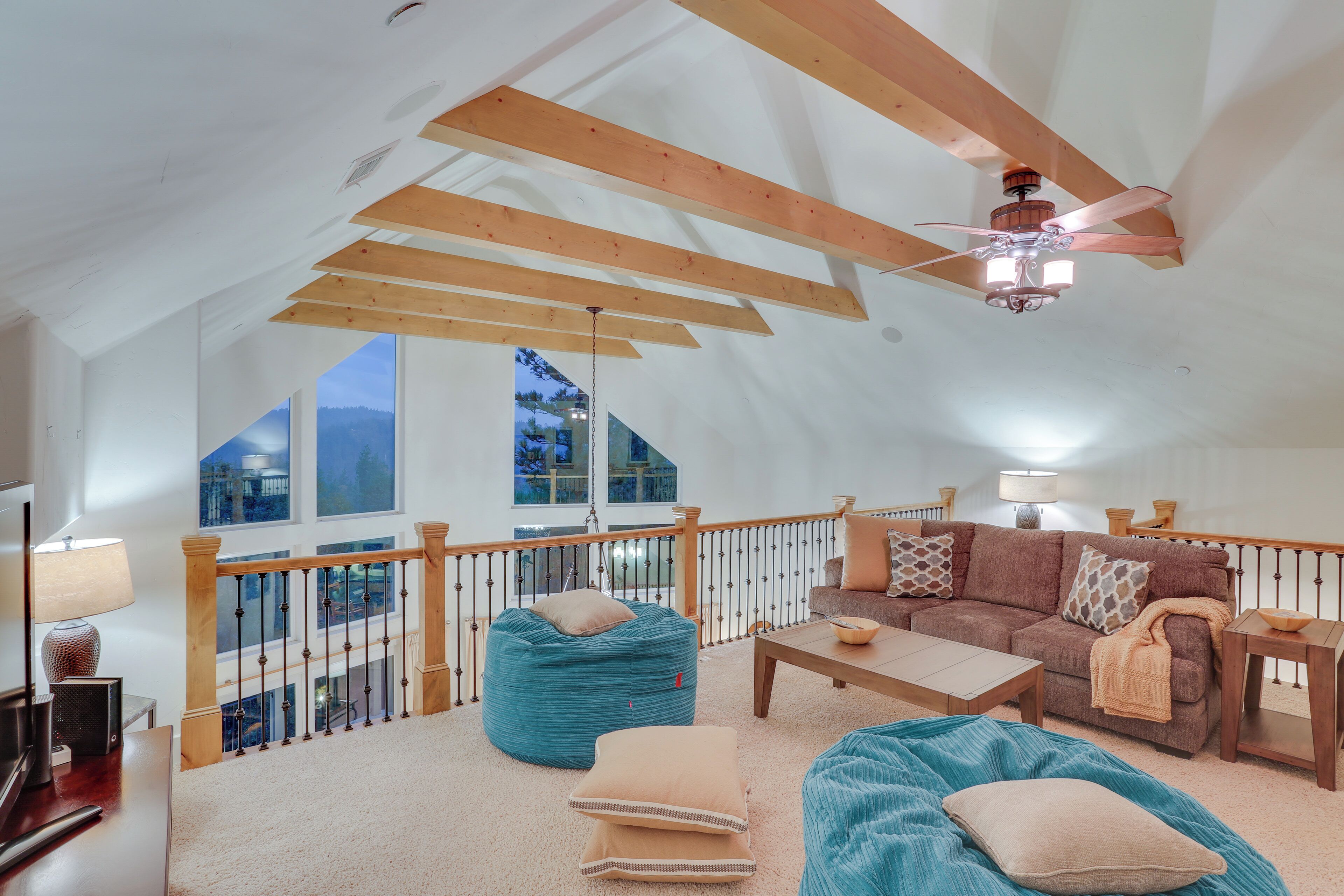 A spacious loft bedroom with a high ceiling, wooden beams, a balcony with a railing, a large window, and a ceiling fan.