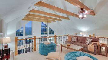 A spacious loft bedroom with a high ceiling, wooden beams, a balcony with a railing, a large window, and a ceiling fan.