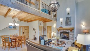 A spacious living area with a high ceiling, wooden beams, a fireplace, and a dining area with wooden furniture. - Beautiful Lake Arrowhead Cabin w/ EV Charger (Lake Arrowhead)