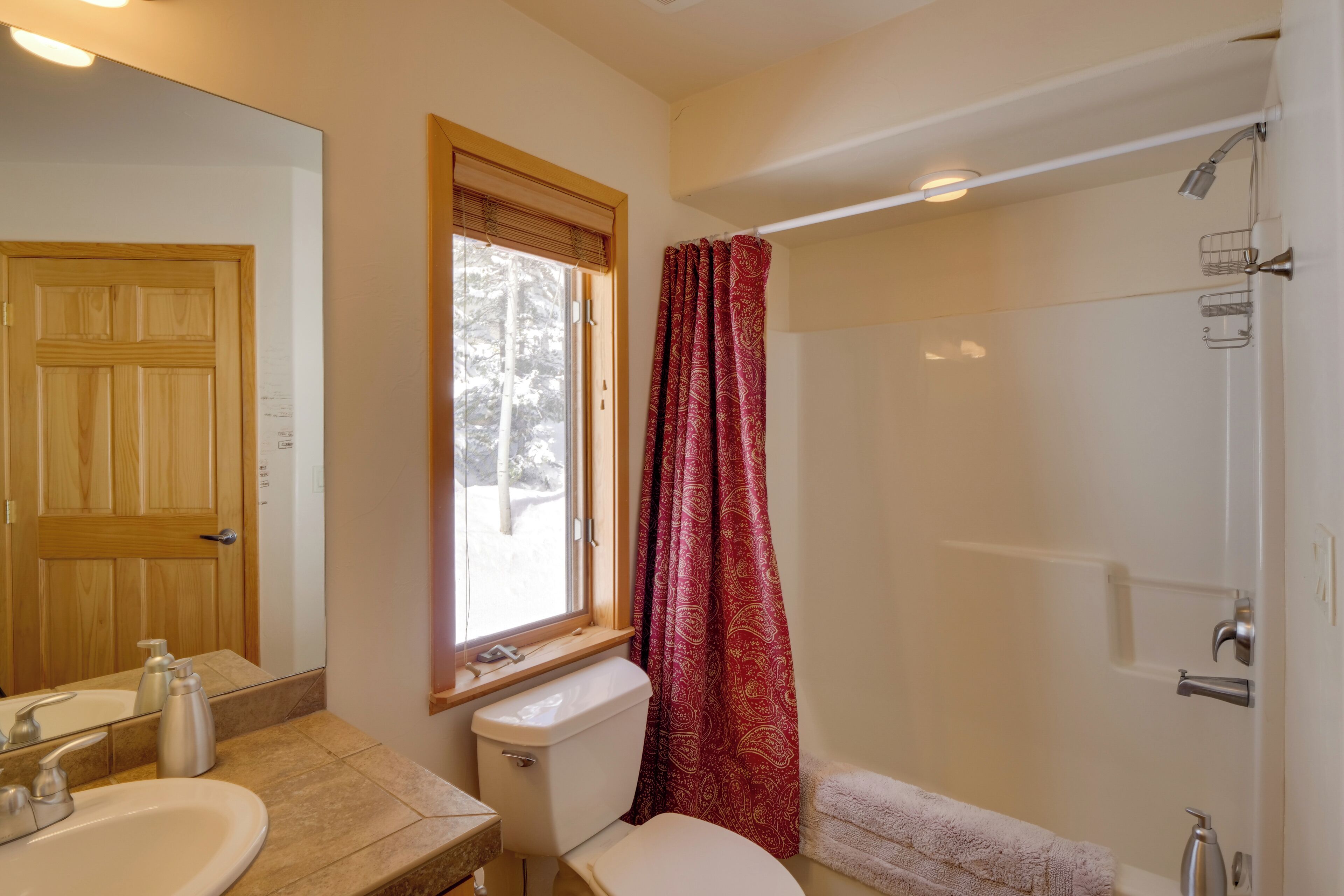 Un baño con puerta de madera, ventana con vistas, inodoro, lavabo y zona de ducha.