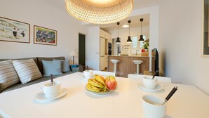 Eine moderne Küche mit weißen Schränken, einem Esstisch mit Obst und Tassen sowie einer hängenden Lampenleuchte.