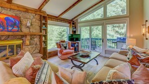 A cozy living room with a stone fireplace, comfortable seating, a television, and large windows offering a view of the outdoors.