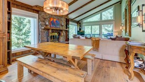 A rustic interior with a wooden dining table, stone fireplace, and large windows offering a view of the outdoors.