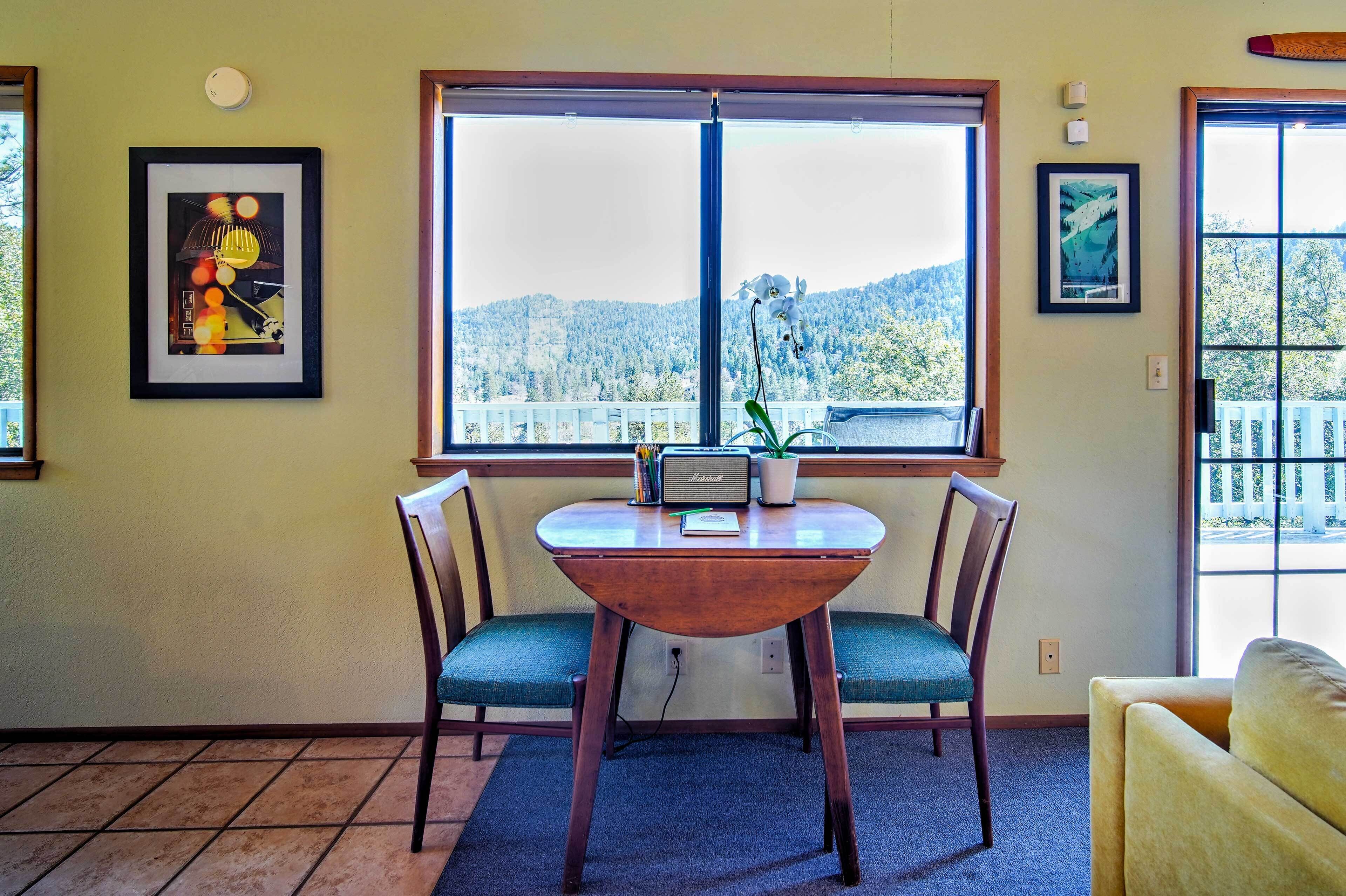 A room with a table and two chairs, a window with a view of a forested area, and two framed pictures on the walls.