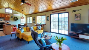 A spacious living room with a yellow sofa, a black recliner, and a TV. There is a kitchen area with wooden cabinets and a dining table. - The Atomic Squirrel Lodge Lake Gregory Getaway! (Crestline)