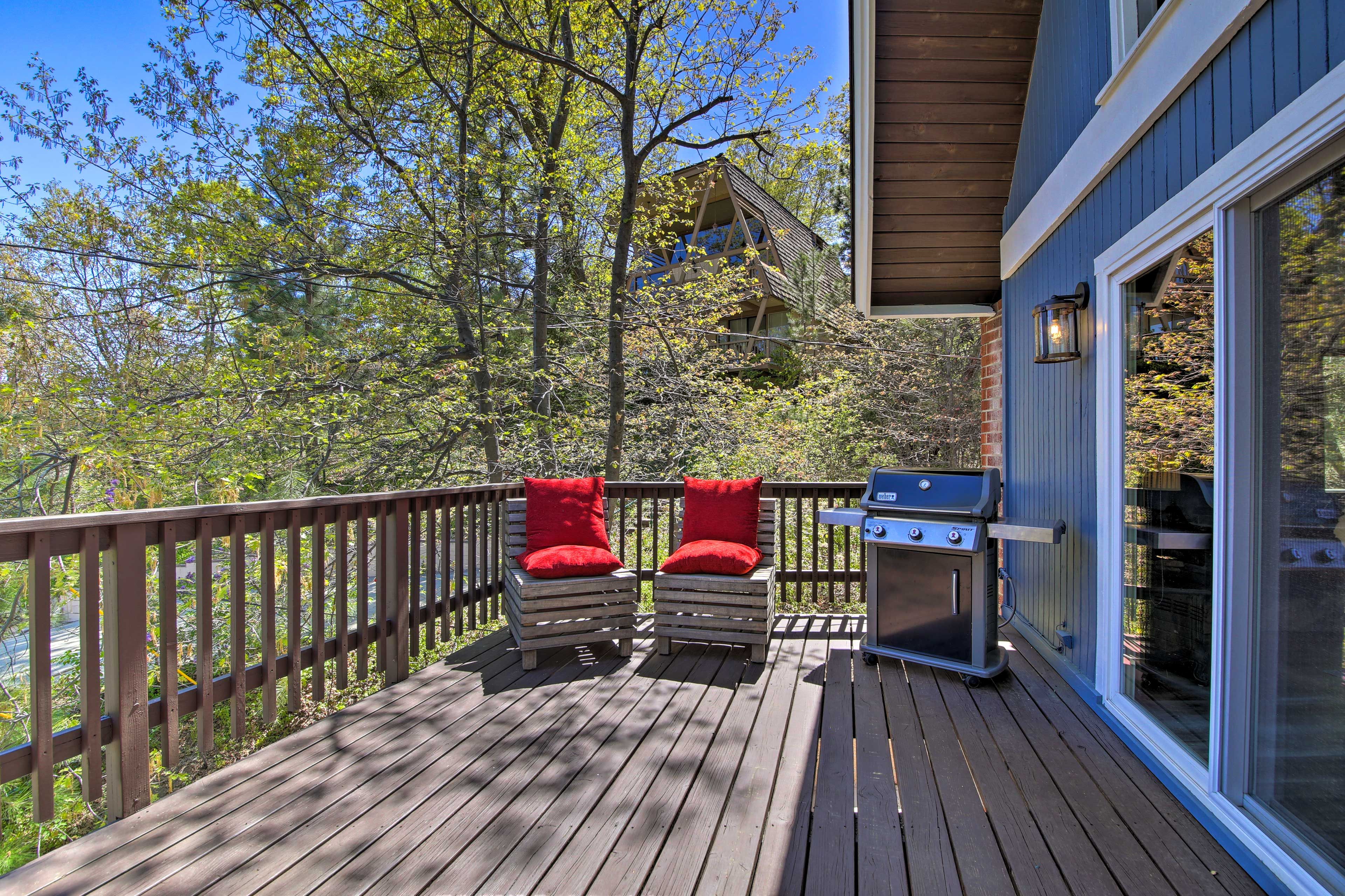 Une terrasse en bois avec deux chaises rouges dotées de coussins, un barbecue et une porte vitrée donnant sur un espace abrité.
