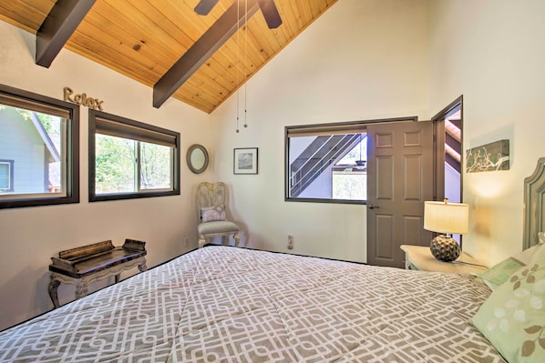 A bedroom with a large bed, a wooden ceiling, a chair, a bedside table with a lamp, and a door leading to a staircase. - Bikes + Game Room: Lovely Lake Arrowhead Home! (Lake Arrowhead)