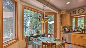 A dining area with a table set for four, wooden chairs, a chandelier, and large windows with a view of trees. - Waterfront Lake Arrowhead Home w/ View & Game Room (Lake Arrowhead)