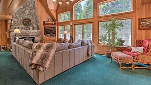 A cozy cabin interior with a stone fireplace, large windows, a sofa, and a dining area. - Waterfront Lake Arrowhead Home w/ View & Game Room (Lake Arrowhead)