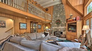 A spacious living room with a large sectional sofa, a stone fireplace, wooden decor, and a balcony with a railing. - Waterfront Lake Arrowhead Home w/ View & Game Room (Lake Arrowhead)