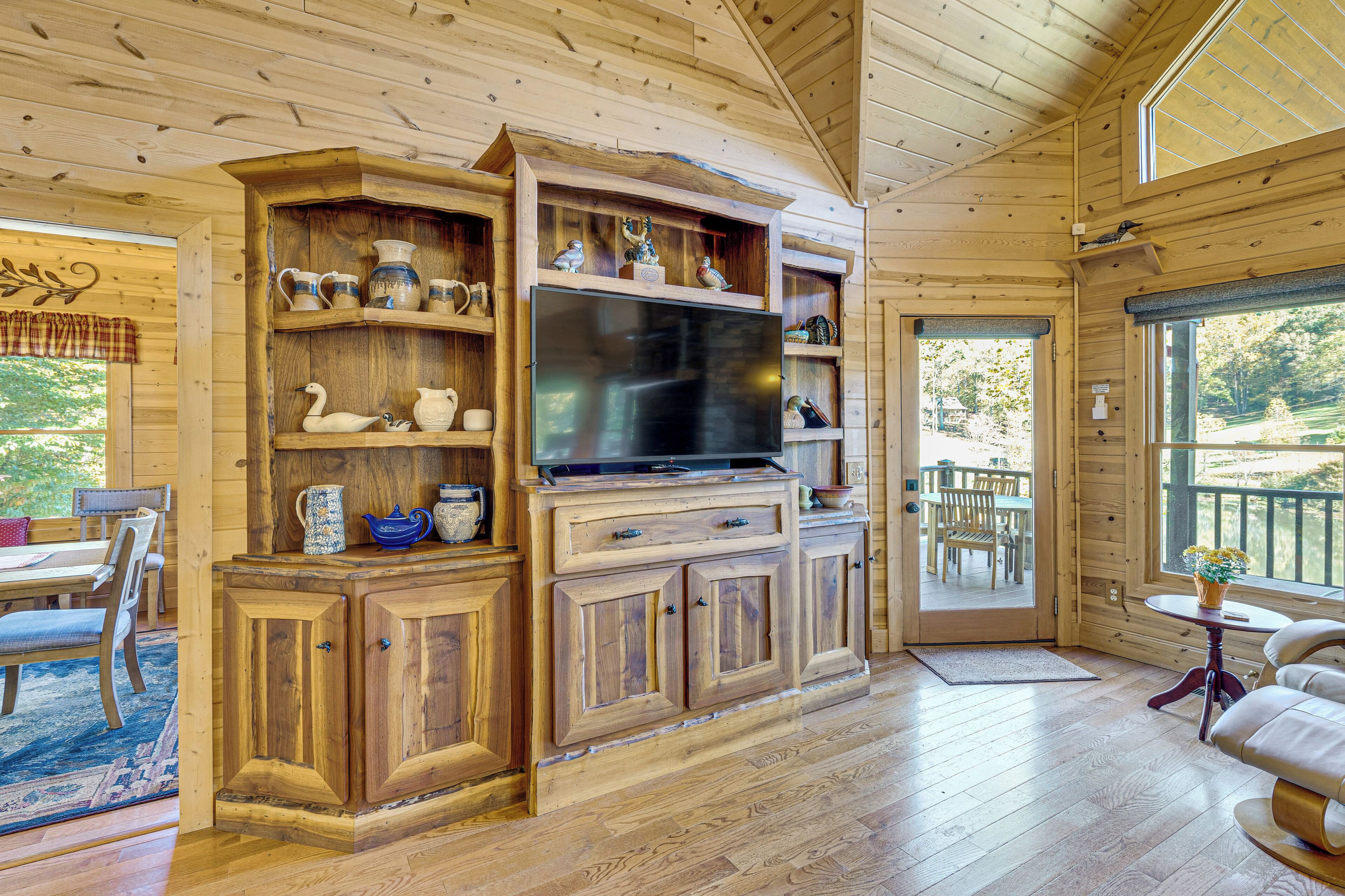 Un intérieur en bois avec un téléviseur posé sur un meuble, des étagères ornées d’objets décoratifs, un coin repas et une vue à travers une porte.