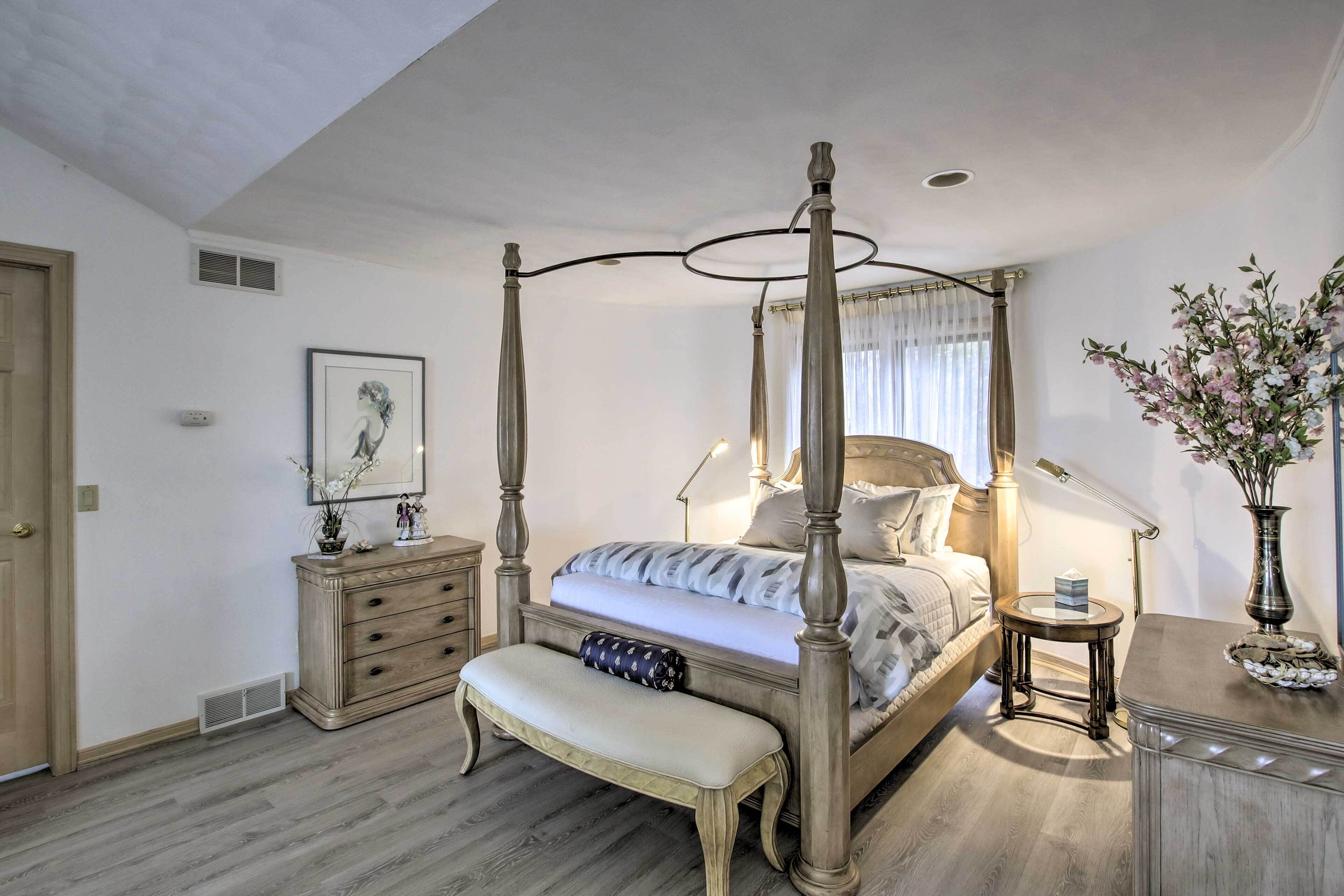 A four-poster bed with a canopy, a bedside table with a lamp, a bench, a dresser, and a vase with flowers.