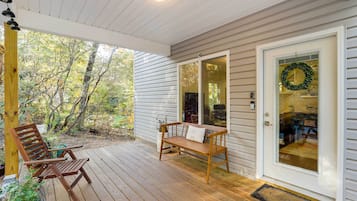 A covered porch with a wooden bench and a chair, a glass door with a wreath, and a view of trees.