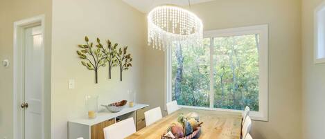 Un comedor con una mesa de madera, sillas blancas, una araña y una ventana con vistas a árboles.