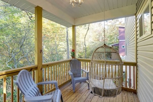 Un porche cubierto con dos sillas Adirondack, una jaula colgante para pájaros y vistas a árboles y una casa.