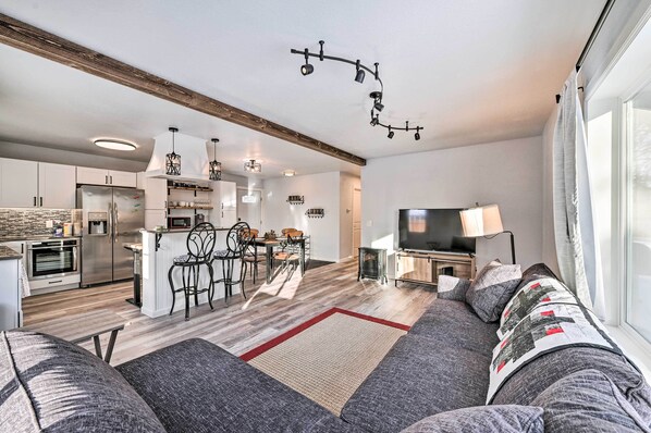 A modern living room with a sofa, a TV, and a dining area with chairs and a table. - Kenai Home w/ Fireplace < 10 Mi to Big Eddy! (Kenai)